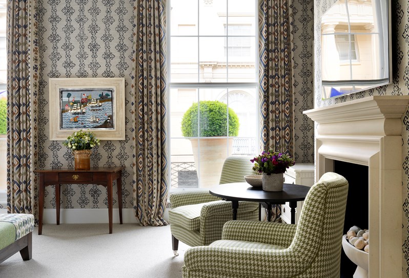 Two large pale green Gingham armchair sit in front of a stone fireplace. In the background are two floor length windows looking out on to the street with a large terracotta pot on the balcony.