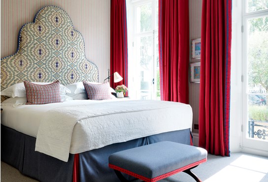 A large spacious bedroom with pale blue and red striped walls and a patterned headboard in a red and white colour. There are two floor to ceiling french windows draped in bright red curtains.