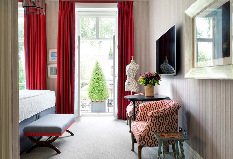 Two small red and white armchairs frame a TV on the side wall. On the back wall are floor to ceiling French windows framed by bright red curtains.