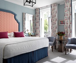 A large spacious bedroom with blue walls and a patterned headboard in a red and white colour. There are two small chairs either side of french windows, with a sunny leafy outlook.
