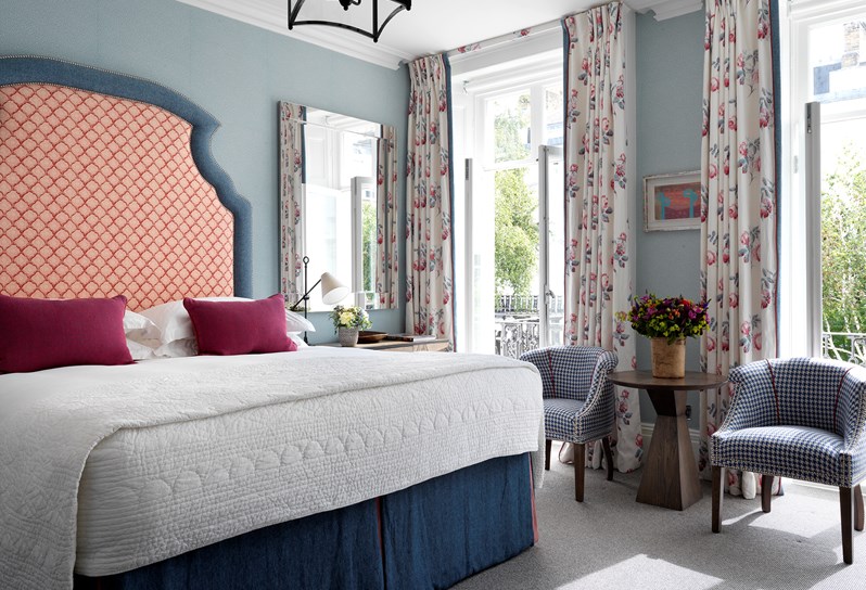 A large spacious bedroom with blue walls and a patterned headboard in a red and white colour. There are two small chairs either side of french windows, with a sunny leafy outlook.