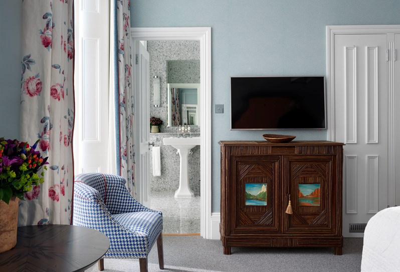 A pale blue bedroom with cream floral curtains. There is an ornate wooden cabinet with a TV. Through an open door you can see through to the en-suite granite bathroom.
