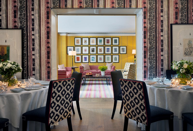 The Covent Garden Hotel Fortune function room with a striped wallpaper and a black table laid for dinner in the foreground. Open double doors look through the bright yellow Lyric room.