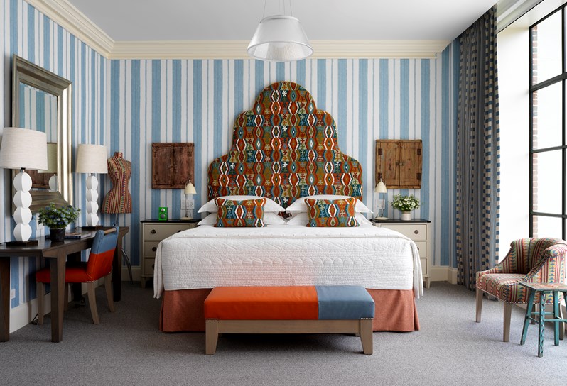 The bedroom room in a one bedroom suite at the Crosby Street Hotel with king size bed with a brown and orange kilim pattern headboard set against blue stripe walls. There are floor length windows with views of the SoHo neighbourhood