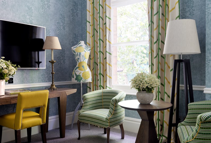 The corner of a hotel bedroom featuring a flat screen television, make up desk and a signature Firmdale mannequin positioned by a window and two armchairs with a table.