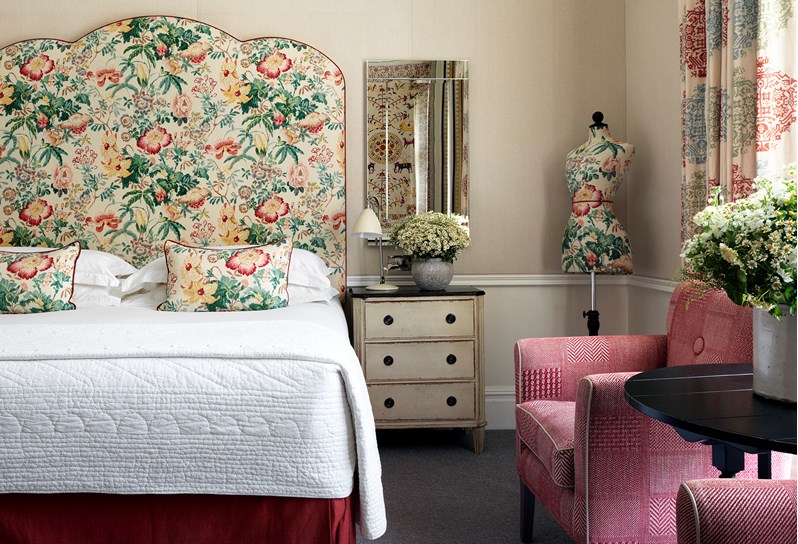 A colourful floral headboard sits above a large bed with a pink armchair to the right hand side and floor-to-ceiling curtains.