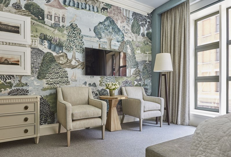 Two cream arm chairs are set against a wall with a hand painted wallpaper depicting green rolling hills with trees and creatures.