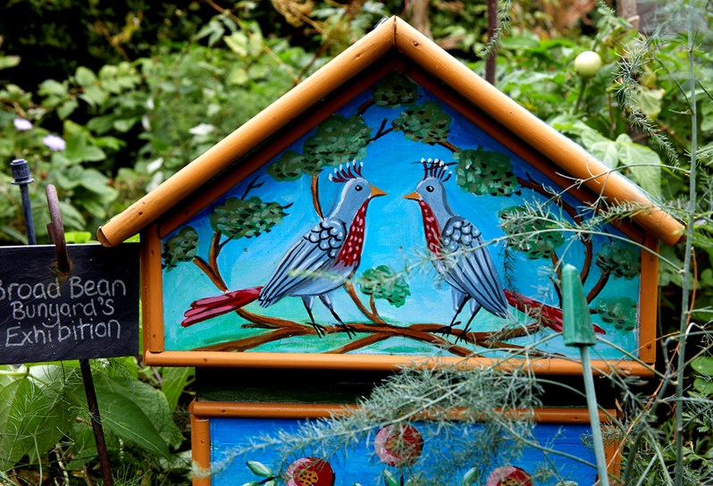 A colourful wooden bird house sits among a garden with a bright blue picture of two birds looking at each other