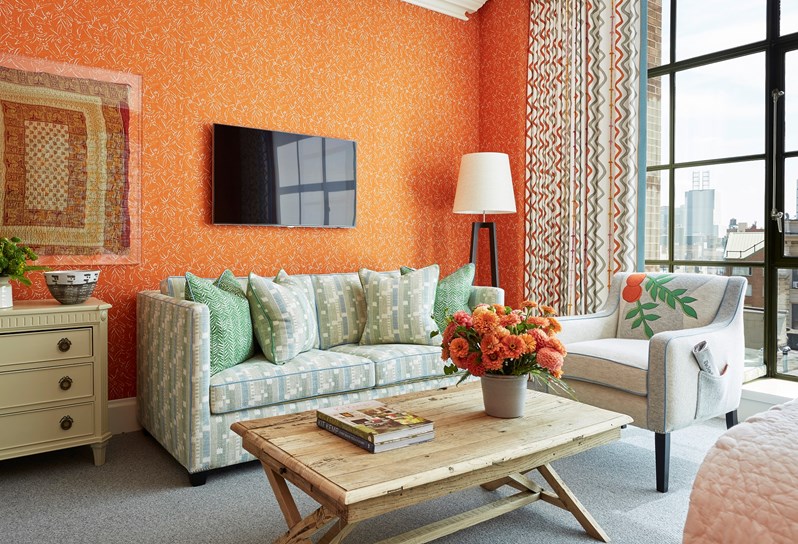 A colourful orange bedroom with a pale green and blue sofa and an arm chair with an embroidered orange set in front of floor to ceiling windows with views of New York