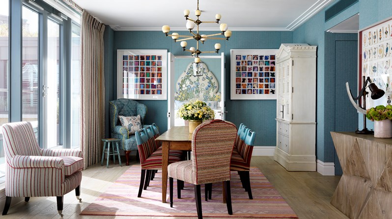 A spacious seating area looking in toward a colorful bedroom, with colorful artwork on each side of the doorway, featuring lots of comfortable seating in two colorful armchairs and heavily patterned sofa places around a wooden coffee table, all with the light from floor to ceiling windows on the left.