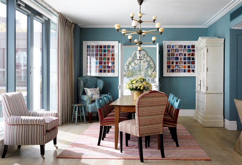 A spacious seating area looking in toward a colorful bedroom, with colorful artwork on each side of the doorway, featuring lots of comfortable seating in two colorful armchairs and heavily patterned sofa places around a wooden coffee table, all with the light from floor to ceiling windows on the left.