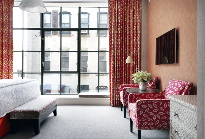 A bedroom with floor to ceiling loft-style windows with two armchairs and flat-screen television mounted to the wall.