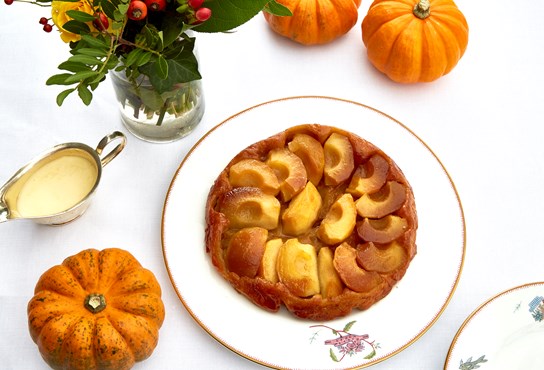 A golden apple start on a Kit Kemp designed plate is surrounded by mini pumpkins, a bouquet of bright flowers and a serving jug of custard.