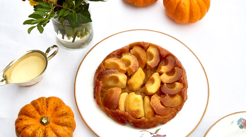 A golden apple start on a Kit Kemp designed plate is surrounded by mini pumpkins, a bouquet of bright flowers and a serving jug of custard.