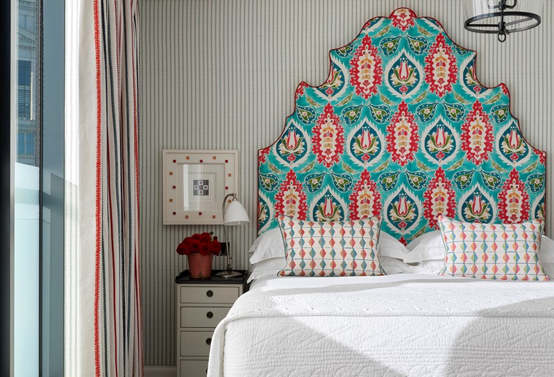 A king-size bed with white linen and a turquoise patterned feature headboard. A cream bed side table with a black top sits to the left hand side of the bed, resting up against an striped fabric wall.