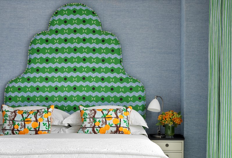 One of three bedrooms in Apartment 22. A king-size bed with green and blue patterned feature headboard rests against a blue fabric wall and a blue footstool rests at the end of the bed. On the left hand side, in front of floor to ceiling windows with balcony doors, are two cream upholstered armchairs with a small wooden table in between.