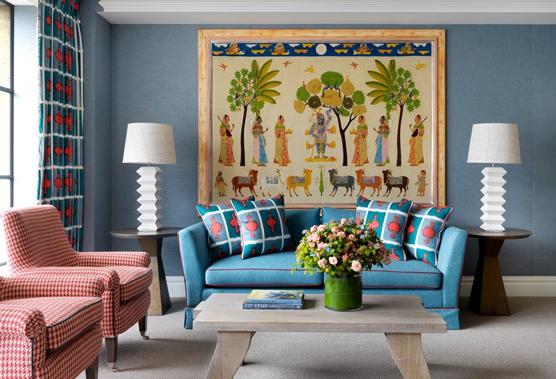 A spacious lounge area in an open plan suite, boasting floor to ceiling windows with a seating arrangement in front; a blue comfortable sofa, a wooden coffee table with fresh flowers, two patterned armchairs and original artwork and quirky design pieces.