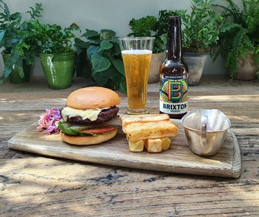 Complete menu shown in front of some plants. Burger, hand cut chips, sauce, on a wooden serving board next to a poured glass of Brixton Pale Ale.