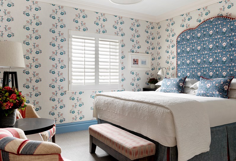 A bright bedroom with a king size bed with a blue headboard and white walls lined in Kit Kemp's Psycho Sprig fabric