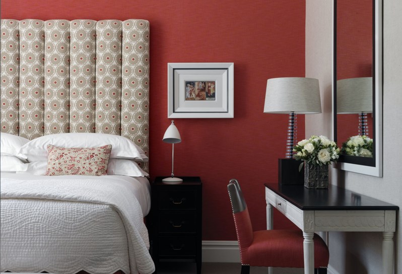 A bright sunny bedroom with red walls and a large statement headboard covered in a cream geometric pattern.