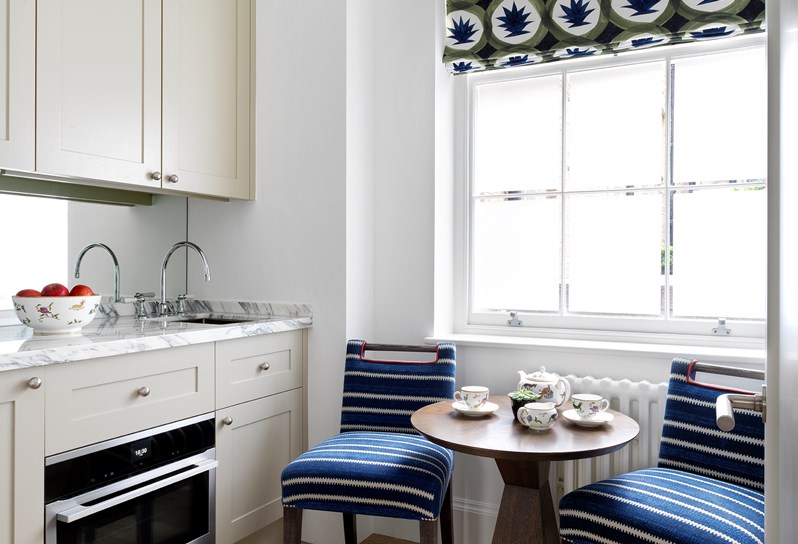 A sunny kitchen with white cabinets and a white marble counter. In front of the window are two blue chairs and a small breakfast table