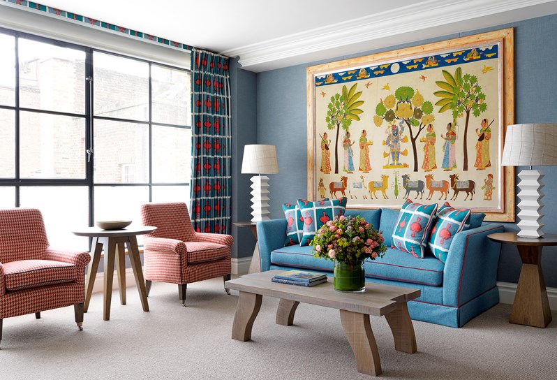 A spacious lounge area in an open plan suite, boasting floor to ceiling windows with a seating arrangement in front; a blue comfortable sofa, a wooden coffee table with fresh flowers, two patterned armchairs and original artwork and quirky design pieces.