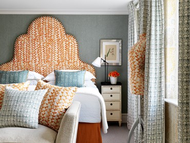 A bedroom and a bed with a large headboard upholstered in an organic floral pattern, a sofa and bedside table.