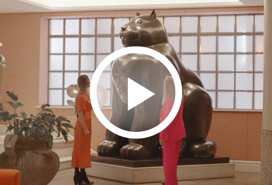 Two woman standing looking up a large Botero sculpture of a cat in the lobby of The Soho Hotel. The image is overlaid with a play button.