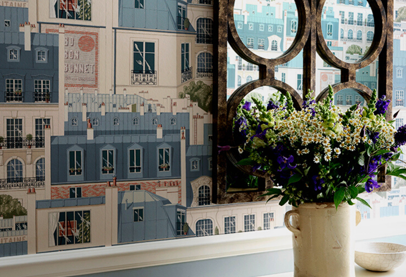 A close up image of decorative wallpaper with buildings printed on it in shades of blue and white. A piece of iron artwork hangs on the wall and there are flowers on a table.