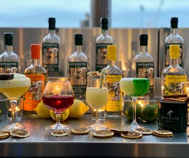 A colourful group of cocktails sit on a silver bar top with bottles of Sipsmith gin behind.