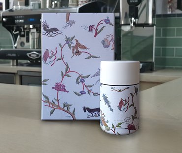 A reusable coffee cup outside a beautiful gift box showing the Kit Kemp Mythical Creatures design on both cup and box in front of a blurred coffee shop background