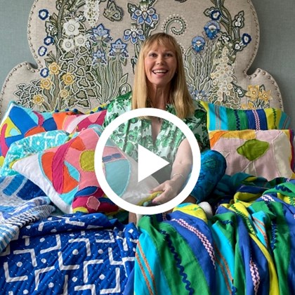 A woman sitting on a bed surrounded by colourful fabrics. The image has a play button overlaid on top.