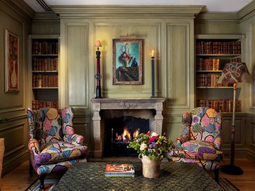 A colourful pair of wingback chairs rest next to a fireplace with an original artwork by the Bloomsbury Group hanging above the fire.