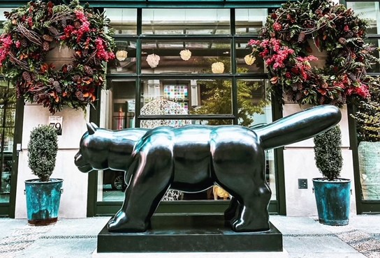 An image of a Fernando Botero sclupture of a cat outside the Crosby Street Hotel. Two large Christmas wreaths hang on either side.