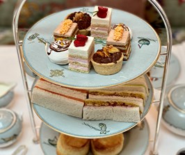 A plate of afternoon tea cakes on a three tier afternoon tea stand. Below are finger sandwiches and scones.