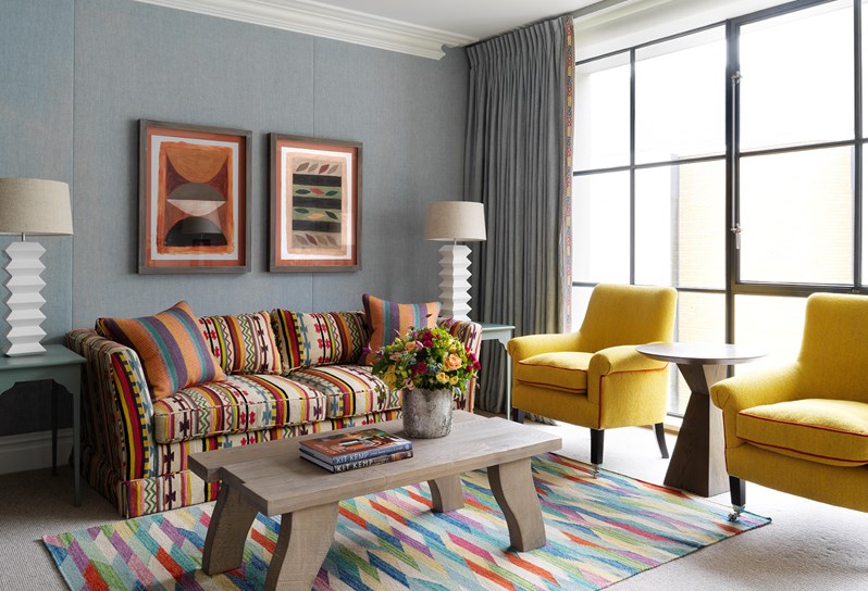 A colourful patterned sofa sitting against a blue linen wall. On the floor under a coffee table is a colourful rug and adjacent to the sofa are floor to ceiling Crittall windows.