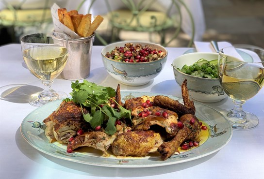 An outdoor table is dressed with a white tablecloth and a selection of dishes including Roast Coronation Chicken as well as two glasses of white wine