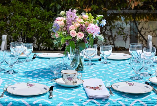 A table covered in a blue patterned tablecloth and laid with kit kemp's mythical creatures china for Wedgwood