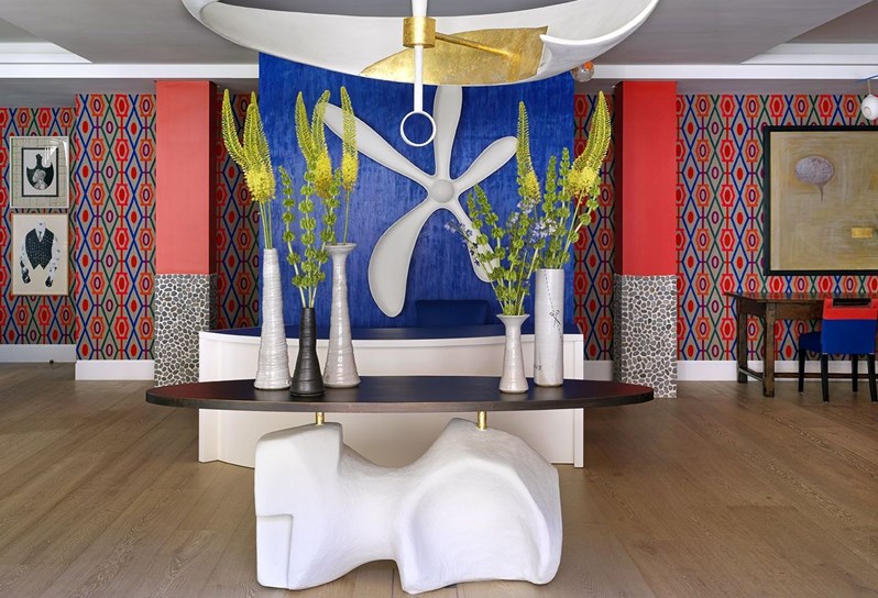 A view of the lobby reception area at 9-11 Richmond Buildings featuring a central table and a large sculptural light hanging above. In the background is a reception desk with a large sculptural flower hung on the blue wall behind. The lobby area is bright and colourful with patterned fabric adorning the walls and statement pieces of artwork.