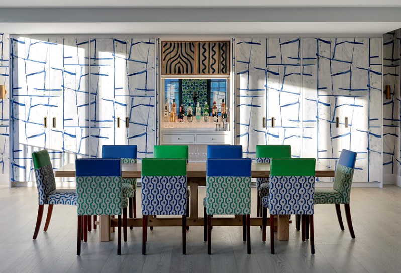 A view of the entertaining area on the top floor at 9-11 Richmond Buildings featuring a large wooden dining table and 10 chairs upholstered in colourful patterned fabrics. Behind the table are floor-to-ceiling cupboards covered in a graphic, geometric print. The central doors of the cupboards are open to reveal a bar area.