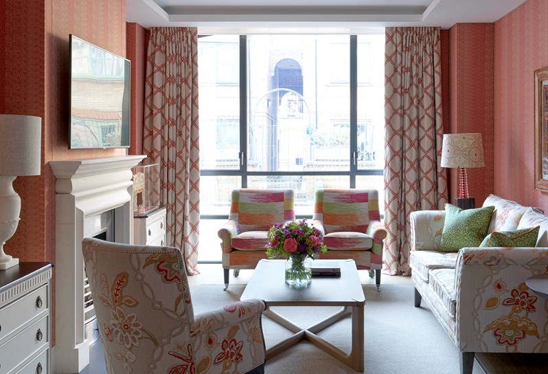 A view of one of the apartments at 9-11 Richmond Buildings. The walls of the living space are upholstered in a soft red fabric and a sofa, two armchairs and a coffee table frame a fireplace on the left hand side of the room. At the end of the room are floor-to-ceiling windows.