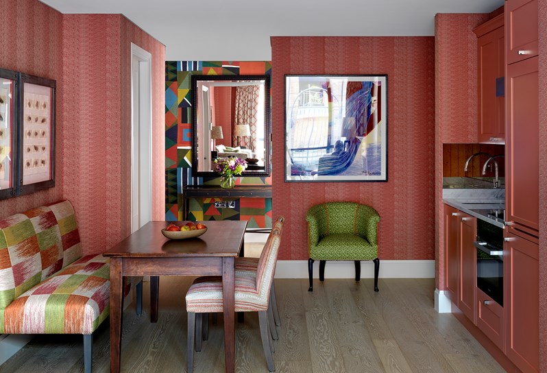 A view of one of the apartments at 9-11 Richmond Buildings. The walls of the living space are upholstered in a soft red fabric, there is a small kitchenette to the right hand side and a dining table with banquette seating and dining chairs to the left.