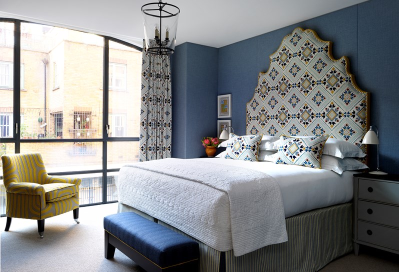 A view of one of the bedrooms in the apartments at 9-11 Richmond Buildings. A large statement headboard and bed dressed in crisp white sheets rest against walls upholstered in a dark navy fabric. There are floor to ceiling windows and a patterned yellow armchair in front of the windows