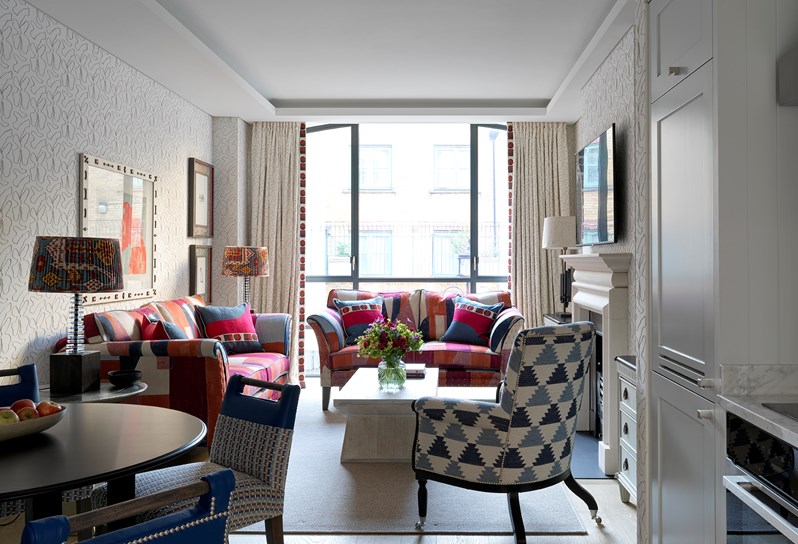 A view of one of the apartments at 9-11 Richmond Buildings. The living space looks through to large floor-to-ceiling windows and features two colourful sofas and a wooden coffee table which frame a fireplace on the right hand wall. In the foreground is a small kitchenette and circular dining table with chairs.