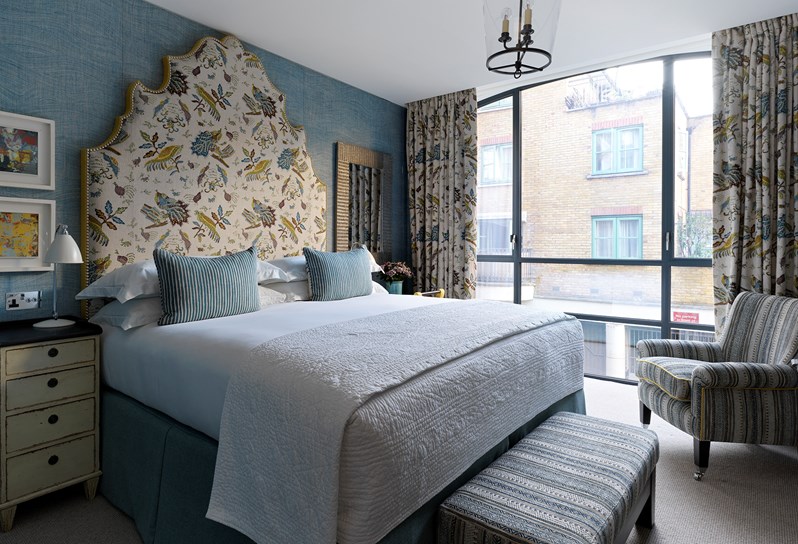A view of one of the bedrooms in the apartments at 9-11 Richmond Buildings. A large statement headboard and bed dressed in crisp white sheets rest against walls upholstered in a soft blue fabric. There are floor to ceiling windows and a patterned armchair in front of the windows.
