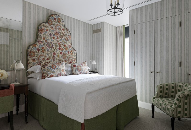 A view of one of the bedrooms in the apartments at 9-11 Richmond Buildings. A large statement headboard and bed dressed in crisp white sheets rest against walls upholstered in a blue and white striped fabric. In the corner of the room is a tub chair upholstered in a green and white fabric.