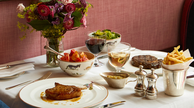 A table dressed with a white tablecloth and vase of flowers with a variety of dishes including a pork chop and side orders including fries
