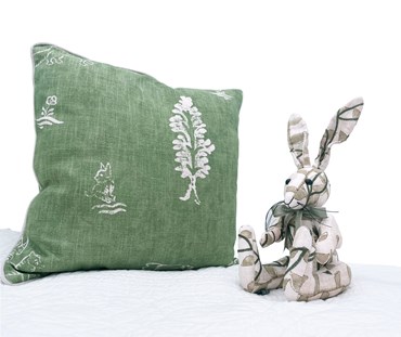 Minnie the rabbit patchwork toy, in "Tea-leaves" fabric : sage green leaves printed and embroidered the outline of dark green leaves on a natural off white linen, with matching ribbon bow around neck, pictured here sitting on a white bedspread , next to a friendly folks basil cushion.