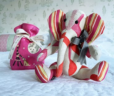 Elephant patchwork toy made out of hot pink, and orange "rik rak" fabric , on a natural background, with hot pink lavender bag, embroidered with moon dog design, (delivered as a gift, in bright pink, and yellow gift box). Presented on a bed with a white bedspread, and a pink and green cushion in the back ground.
