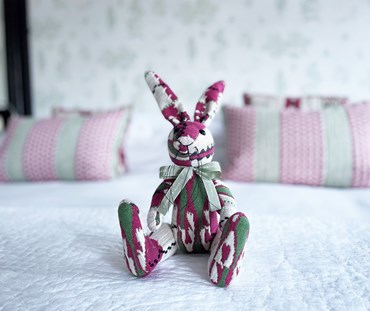 The Minnie "Chubby check " bunny is a new and limited edition member of Kit's patchwork Toy collection. Made from Kit's "Chubby Check " Woven Cotton fabric, in Hot Pink Green and Ecru, inspired by a blanket. This toy is pictured sat on a bed.